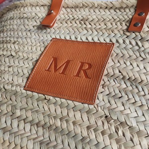 Puede incluir: Un bolso de paja tejido con un parche de cuero marr&oacute;n en el que est&aacute;n bordadas las iniciales "MR" en blanco.