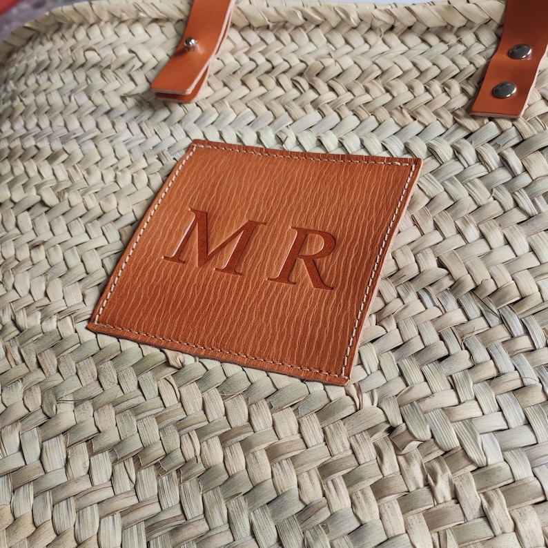 Puede incluir: Un bolso de paja tejido con un parche de cuero marr&oacute;n en el que est&aacute;n bordadas las iniciales "MR" en blanco.