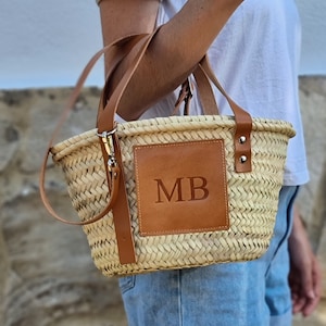 May include: Woven straw handbag with brown leather accents. Features a rectangular leather patch with the initials "MB". Includes a detachable shoulder strap and two short handles. The bag is held by a person wearing a white shirt and denim shorts.