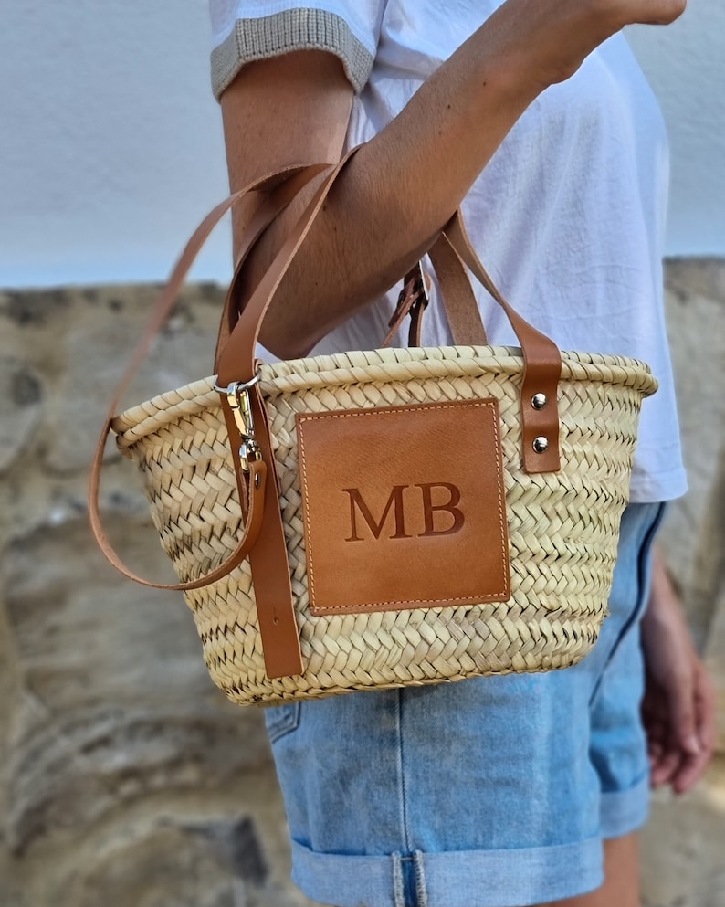 May include: Woven straw handbag with brown leather accents. Features a rectangular leather patch with the initials "MB". Includes a detachable shoulder strap and two short handles. The bag is held by a person wearing a white shirt and denim shorts.