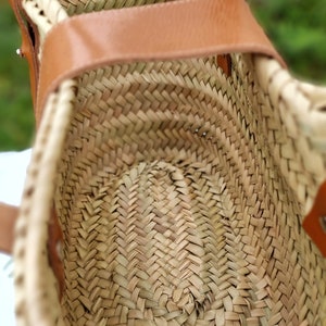 May include: A woven straw bag with a brown leather handle. The bag is open and the inside is visible. The straw is woven in a herringbone pattern.