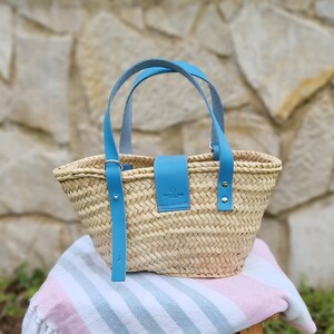 May include: A woven straw tote bag with blue leather handles and a blue leather strap across the front. The bag has a small leather tag with the text "The Carry Shop" on it.