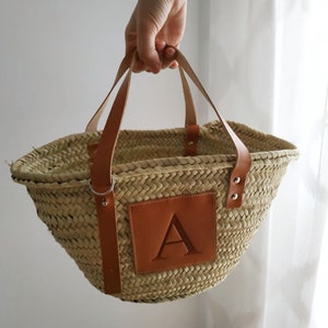 Puede incluir: Un bolso tote de paja natural tejido con asas de cuero y un parche de cuero marr&oacute;n con la letra "A" en la parte delantera.
