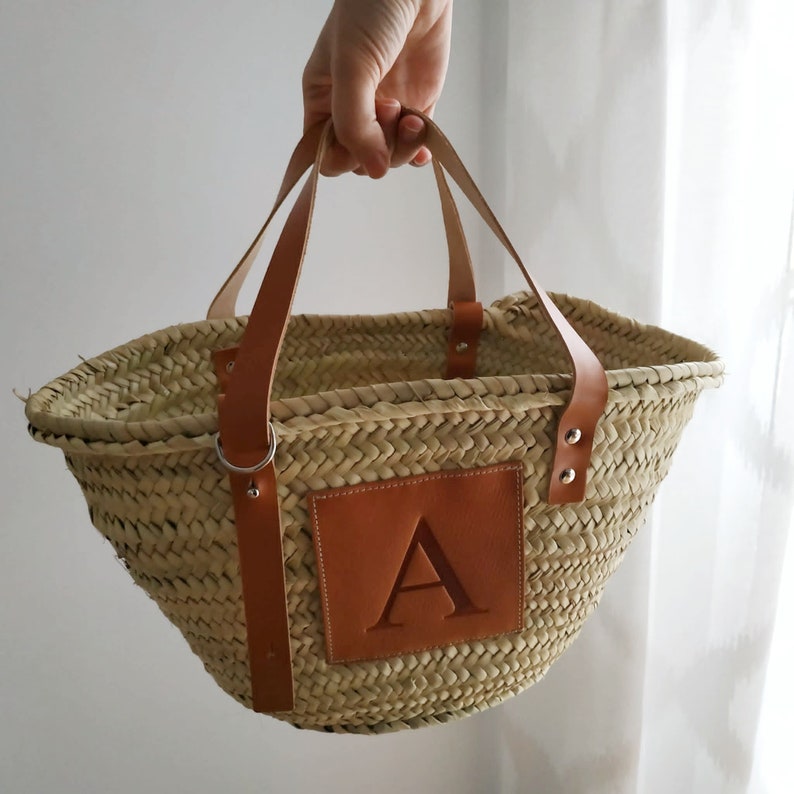 Puede incluir: Un bolso tote de paja natural tejido con asas de cuero y un parche de cuero marr&oacute;n con la letra "A" en la parte delantera.