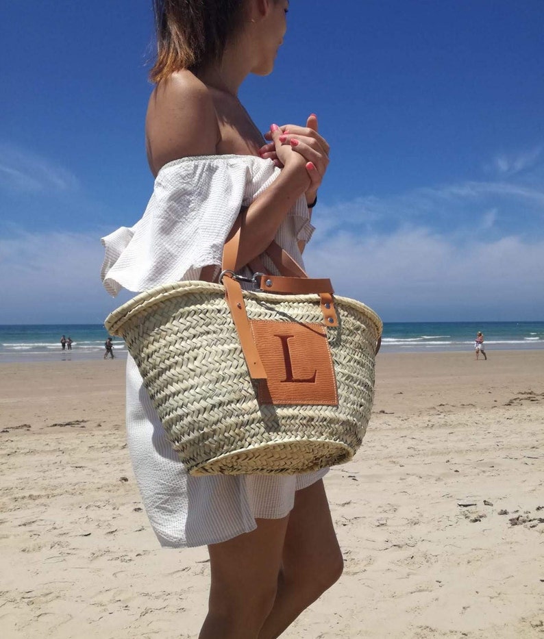 Puede incluir: Una mujer en una playa llevando un gran bolso de mimbre con asa de cuero y un parche de cuero con la letra "L" en &eacute;l.