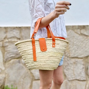 May include: A woven straw tote bag with orange leather handles and a leather patch with the letter "A" on the front. The bag is being held by a person wearing a white dress.