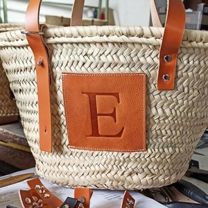 Puede incluir: Un bolso de mimbre tejido con asas de cuero y un parche de cuero con la letra "E" en relieve. El bolso est&aacute; sobre una mesa con algunas correas de cuero y unas tijeras.