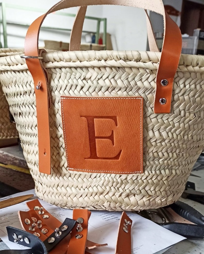 Puede incluir: Un bolso de mimbre tejido con asas de cuero y un parche de cuero con la letra "E" en relieve. El bolso est&aacute; sobre una mesa con algunas correas de cuero y unas tijeras.