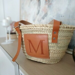 Puede incluir: Un bolso de mimbre tejido con asa de cuero marr&oacute;n y un parche de cuero marr&oacute;n con la letra "M" en la parte delantera.