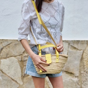 May include: A woman is wearing a white eyelet blouse and denim shorts. She is carrying a straw tote bag with a yellow leather strap and handle.
