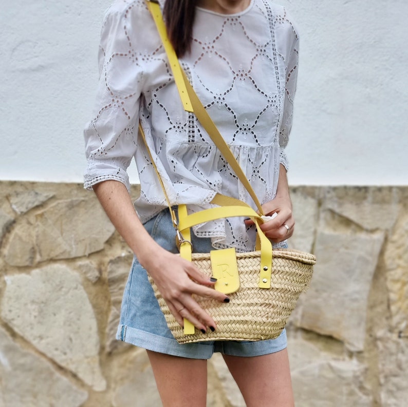 May include: A woman is wearing a white eyelet blouse and denim shorts. She is carrying a straw tote bag with a yellow leather strap and handle.