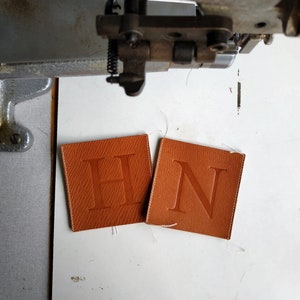 Puede incluir: Dos cuadrados de cuero marr&oacute;n con las letras "H" y "N" en relieve. Los cuadrados est&aacute;n cosidos en los bordes.