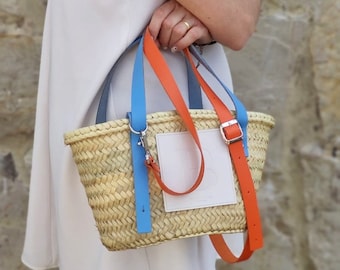 Peut inclure: Un sac cabas en paille tressée avec des sangles bleues et orange. Le sac a un patch blanc sur le devant.