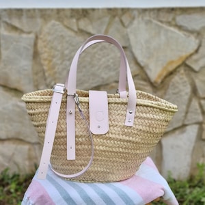 May include: A light pink woven straw tote bag with leather handles and a leather strap. The bag has a leather patch with the letter "C" on the front.