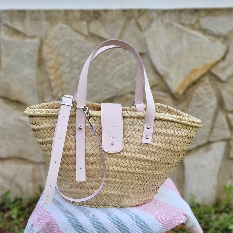 May include: A light pink woven straw tote bag with leather handles and a leather strap. The bag has a leather patch with the letter "C" on the front.