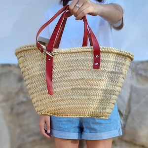 May include: A woven straw tote bag with red leather handles. The bag has a rectangular shape with a wide opening. The red handles are attached with silver-colored hardware. The bag is held up by a person.