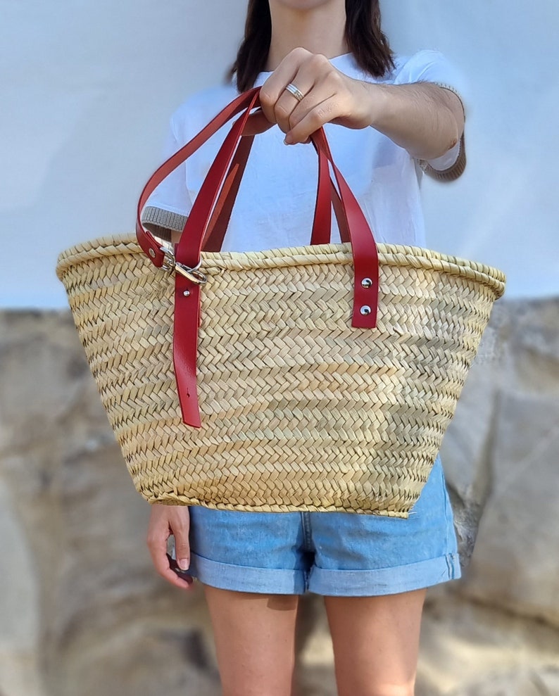 May include: A woven straw tote bag with red leather handles. The bag has a rectangular shape with a wide opening. The red handles are attached with silver-colored hardware. The bag is held up by a person.
