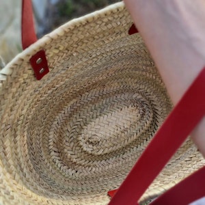May include: Woven straw tote bag with red leather handles and accents. The bag has an oval shape and a natural, light brown color. The red handles are attached with rivets. The bag is designed for carrying items.