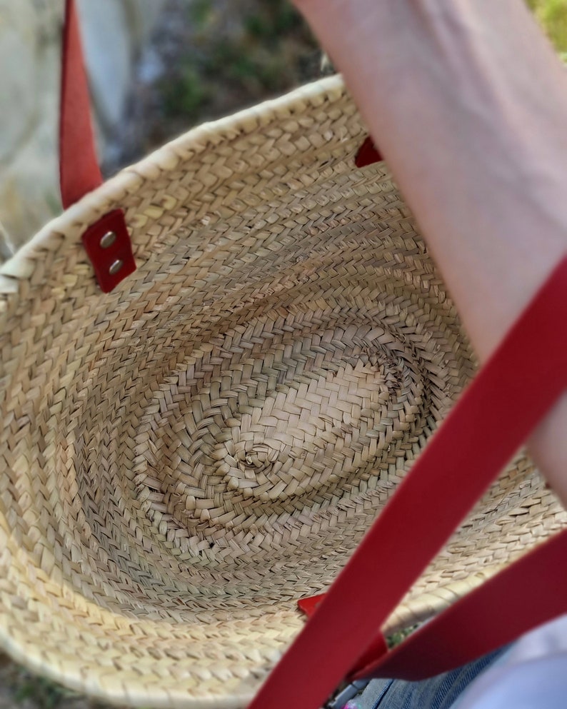 May include: Woven straw tote bag with red leather handles and accents. The bag has an oval shape and a natural, light brown color. The red handles are attached with rivets. The bag is designed for carrying items.
