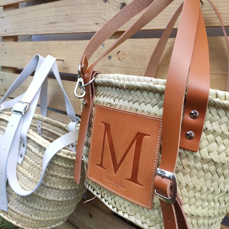Puede incluir: Un bolso tote de cuero marr&oacute;n con un cuerpo de paja tejido. El bolso tiene un parche de cuero grande con la letra "M" y el texto "The Color Bags" en la parte delantera.