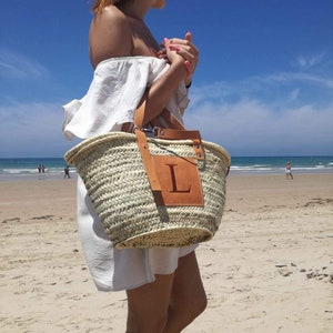 May include: A woven straw tote bag with a leather patch featuring the letter "L". The bag has brown leather handles and is being carried on a sandy beach under a bright blue sky. The bag is a light tan color.