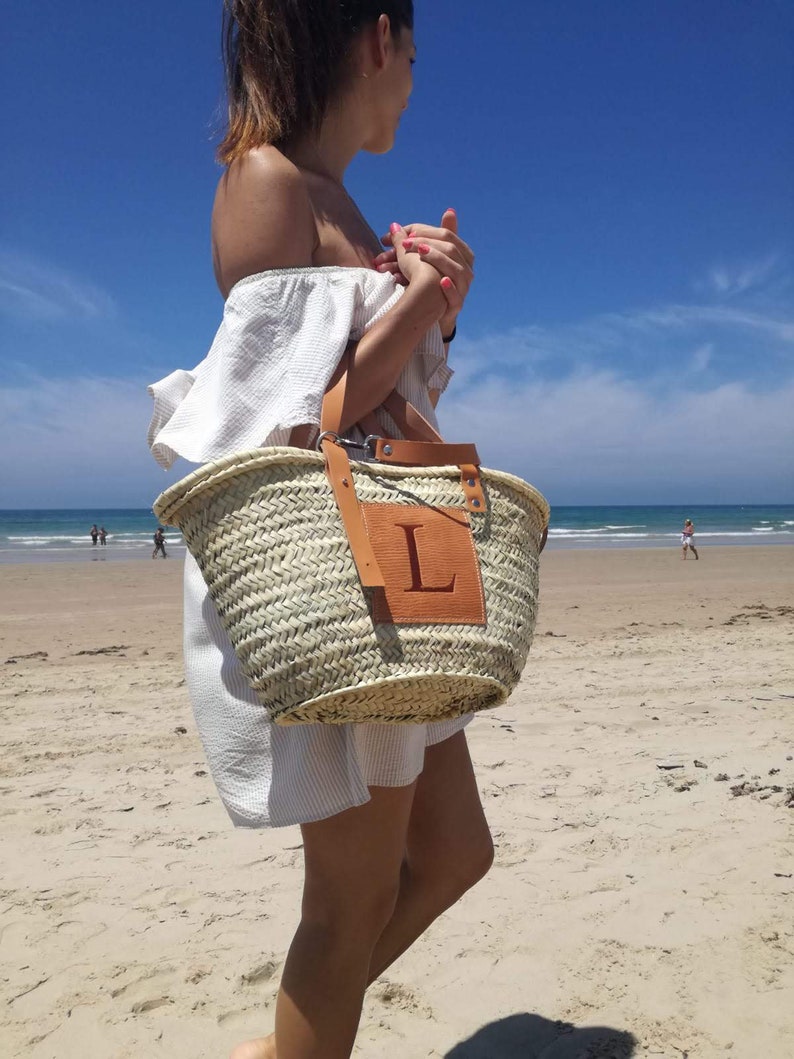 May include: A woven straw tote bag with a leather patch featuring the letter "L". The bag has brown leather handles and is being carried on a sandy beach under a bright blue sky. The bag is a light tan color.