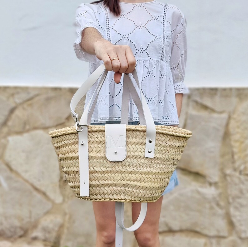 May include: A white leather-trimmed straw tote bag with a white leather initial "M" on the front. The bag has a long white strap that can be worn over the shoulder.