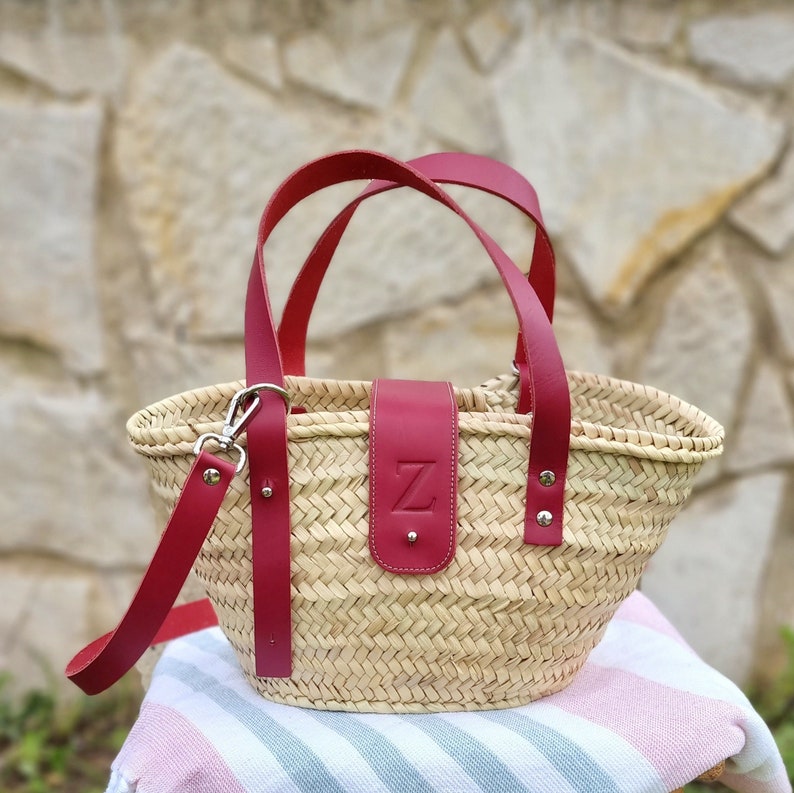 May include: A woven straw tote bag with red leather handles and a red leather strap. The bag has a red leather flap with the letter "Z" embossed on it.