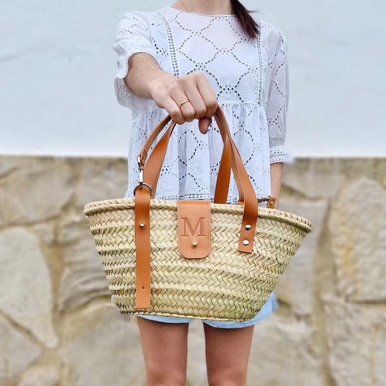 May include: A straw tote bag with brown leather straps and a leather patch with the letter "M" on the front.
