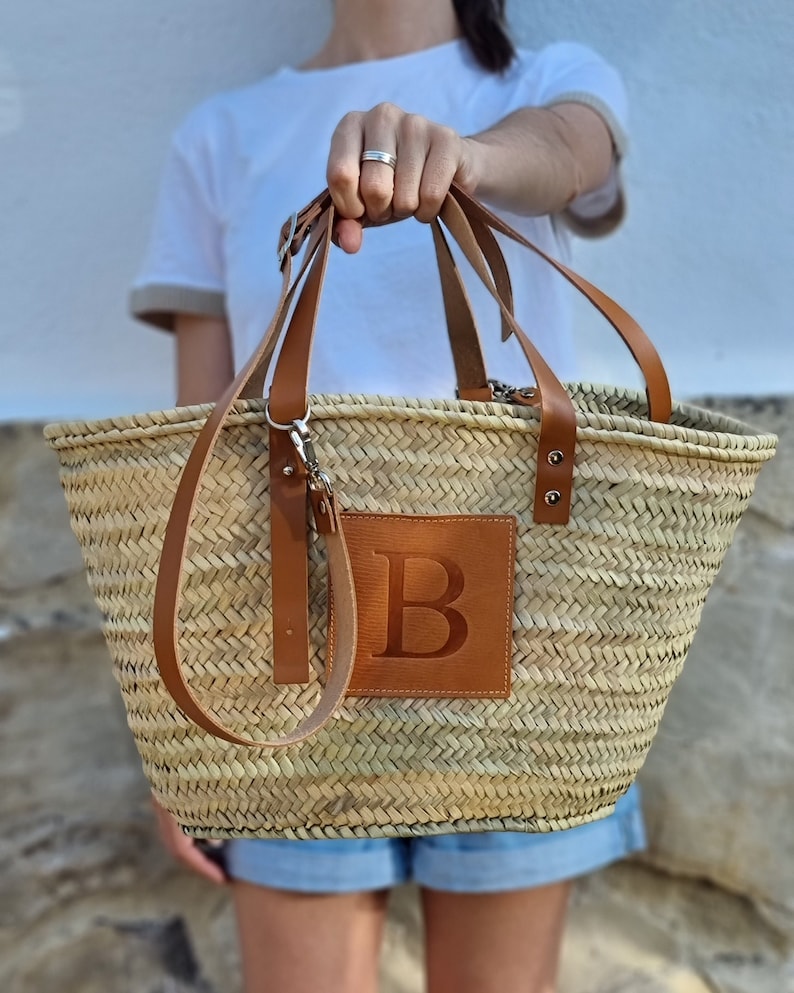 May include: Woven straw tote bag with brown leather handles and a leather patch with the letter "B". The bag has a rectangular shape and is held by a person. The bag is a natural color.