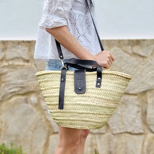 May include: A large, woven straw tote bag with black leather handles and a black leather patch with the letter "B" on the front. The bag is being held by a person wearing a white blouse and denim shorts.