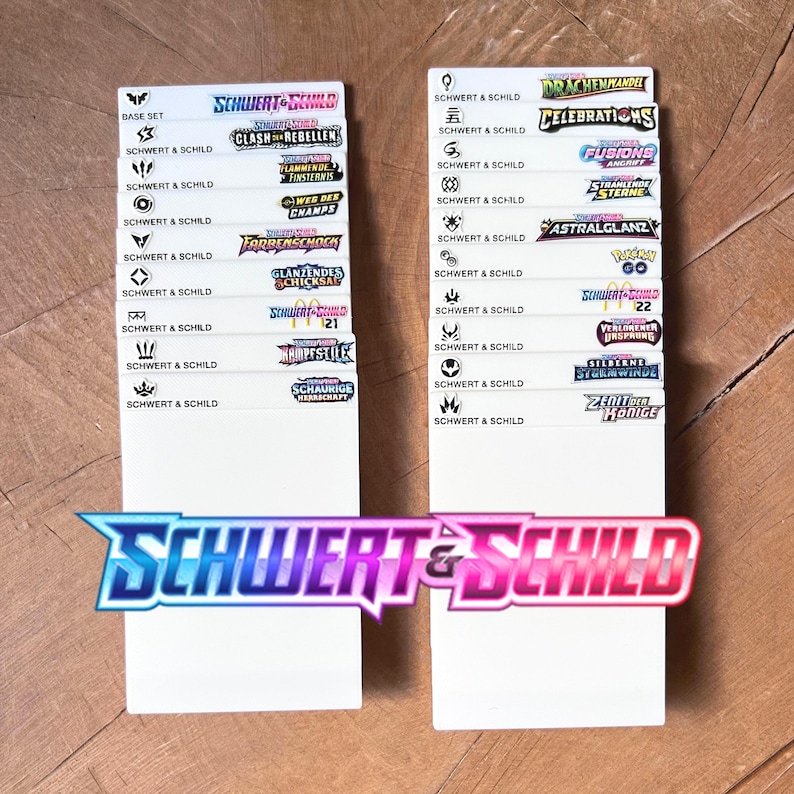 Peut inclure: Deux s&eacute;parateurs rectangulaires blancs pour cartes &agrave; collectionner, chacun pr&eacute;sentant une colonne de logos imprim&eacute;s et le texte Schwert & Schild. Les logos repr&eacute;sentent diverses extensions, dont Base Set, Clash der Rebellen, et d'autres, avec le logo Schwert & Schild en bas.