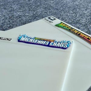 Peut inclure: Deux cartes rectangulaires blanches en plastique avec des surfaces textur&eacute;es. Une carte pr&eacute;sente le texte bleu et blanc WACHSENDES CHAOS avec un graphique d'&eacute;claboussure d'eau. L'autre carte affiche le texte MEGA-ENTWICKLUNG et BASE SET en noir.