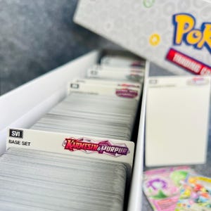 Peut inclure: Une bo&icirc;te de rangement en carton blanc remplie d'une grande pile de cartes &agrave; collectionner. La bo&icirc;te porte le texte Pokemon Trading Card Game. Des s&eacute;parateurs sont visibles, marqu&eacute;s SVI Base Set et le logo Karmesin & Purpur.