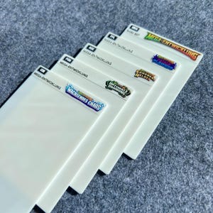 Peut inclure: Un ensemble de cinq s&eacute;parateurs rectangulaires blancs en plastique pour cartes &agrave; collectionner, chacun dot&eacute; d'un onglet noir avec un code &agrave; trois lettres et le texte MEGA-ENTWICKLUNG. Les onglets pr&eacute;sentent des logos color&eacute;s : WACHSENDES CHAOS, OPTIMALE ORDNUNG, ERHABENE HELDEN, FATALE PR&Auml;ZISION et BASE SET.