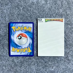 Peut inclure: Une carte &agrave; collectionner rectangulaire pr&eacute;sentant le logo Pok&eacute;mon bleu et blanc avec une Pok&eacute; Ball centrale, plac&eacute;e &agrave; c&ocirc;t&eacute; d'un &eacute;tui &agrave; cartes rectangulaire blanc portant le texte MEG BASE SET et MEGA-ENTWICKLUNG sur le dessus.