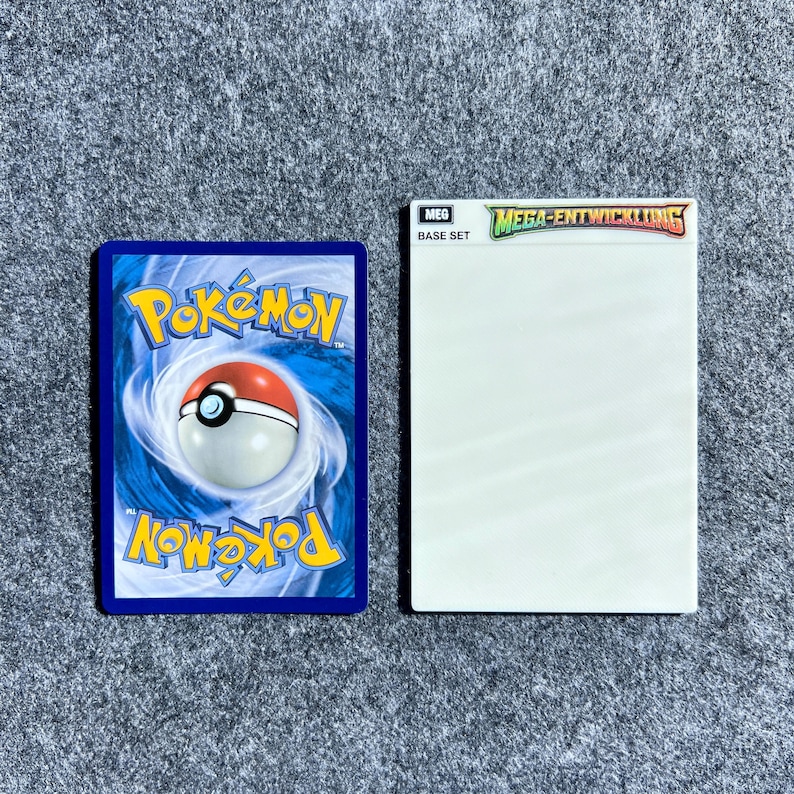 Peut inclure: Une carte &agrave; collectionner rectangulaire pr&eacute;sentant le logo Pok&eacute;mon bleu et blanc avec une Pok&eacute; Ball centrale, plac&eacute;e &agrave; c&ocirc;t&eacute; d'un &eacute;tui &agrave; cartes rectangulaire blanc portant le texte MEG BASE SET et MEGA-ENTWICKLUNG sur le dessus.