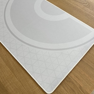 May include: A light gray mousepad with a geometric design. The design features a semi-circular pattern with concentric lines and a triangular pattern in the lower section. The mousepad has a white border and is on a wooden surface.