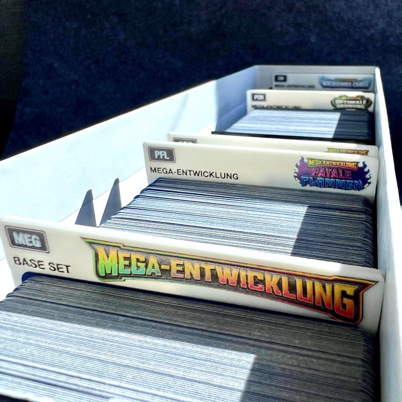 Peut inclure: Une bo&icirc;te de rangement blanche contenant plusieurs piles de cartes &agrave; collectionner organis&eacute;es par des s&eacute;parateurs en plastique blanc. Chaque s&eacute;parateur comporte des &eacute;tiquettes imprim&eacute;es, dont "MEG BASE SET" et "MEGA-ENTWICKLUNG FATALE FLAMMEN", avec du texte et des logos color&eacute;s.