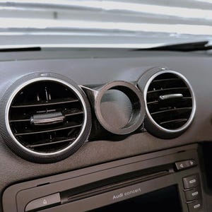 Puede incluir: Un salpicadero de coche negro con dos rejillas de ventilación circulares y un indicador central. Las rejillas de ventilación tienen un borde circular texturizado. Debajo se encuentra un equipo de música con el texto "Audi concert".