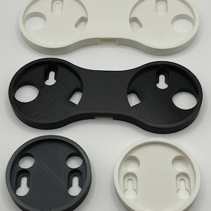 May include: A selection of 3D-printed wall mounts in black and white. The mounts have circular and oblong shapes with holes and keyhole slots for mounting. Designs include single and double-mount configurations, suitable for various applications.