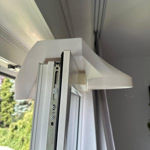 May include: A white 3D-printed plastic ventilation hood attachment mounted onto the top frame of a white window. The attachment features a wide, angled canopy design that extends outward from the window's metal locking mechanism and frame.
