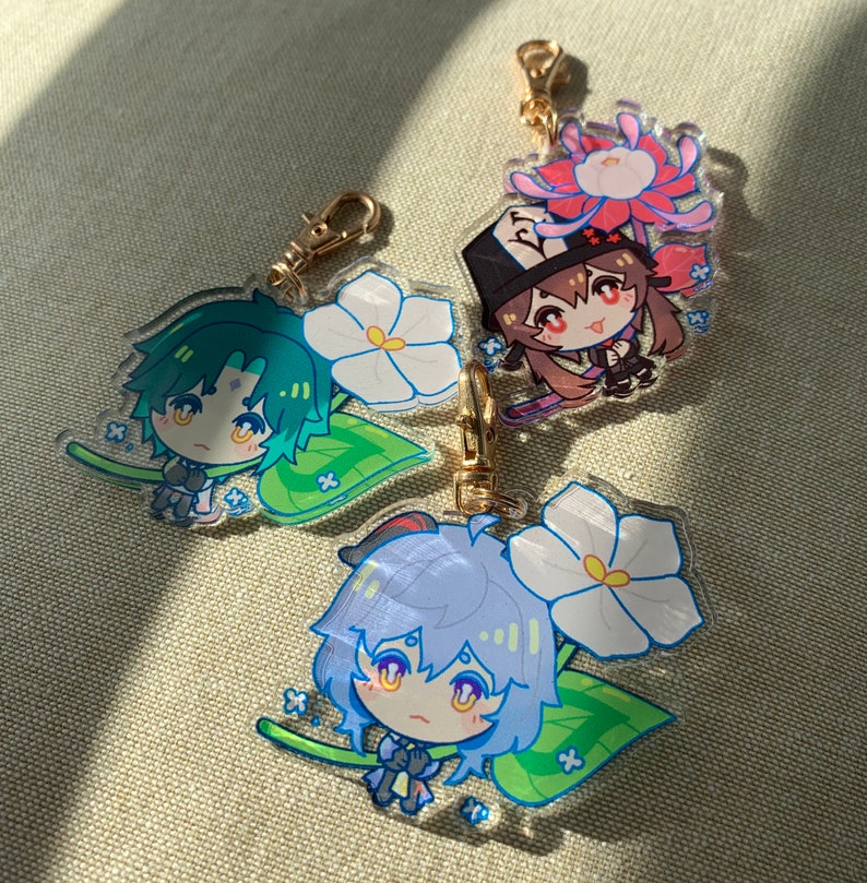 Puede incluir: Tres llaveros de acr&iacute;lico transparente con personajes de estilo anime con flores. El primer llavero tiene un personaje de pelo verde con una flor blanca. El segundo llavero tiene un personaje de pelo marr&oacute;n con una flor rosa. El tercer llavero tiene un personaje de pelo azul con una flor blanca.