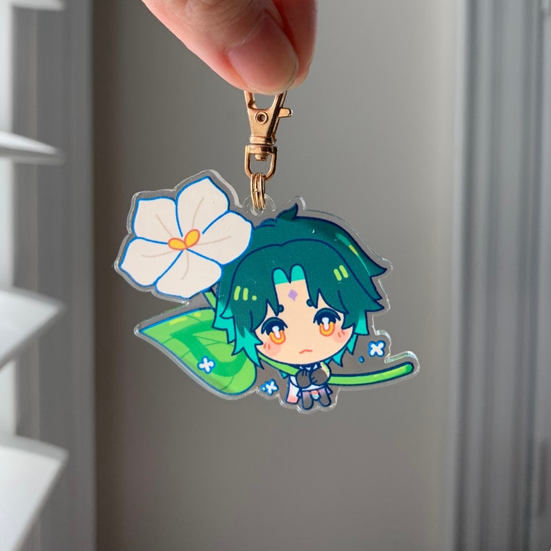 Puede incluir: Un personaje de anime de pelo verde con una flor blanca y hojas verdes, sosteniendo un peque&ntilde;o tallo verde. El personaje lleva un atuendo azul claro y rosa y un llavero dorado est&aacute; unido en la parte superior.