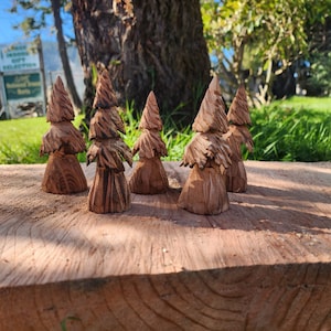 May include: Six small wooden carvings of pine trees, arranged in a row on a wooden surface. The carvings are a light brown color and have a rustic, hand-crafted look.