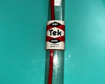 May include: A red toothbrush with white bristles enclosed in a clear cylindrical plastic case with red caps. A white paper label wraps around the center, featuring the text 33c, Tek, HARD, and TEK-HUGHES Watervliet, N.Y. in black and red ink.