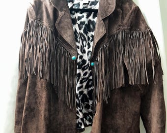 May include: A brown suede jacket featuring long fringe detailing across the chest and sleeves. The jacket includes two turquoise-colored circular buttons at the front closure and is lined with a black and white leopard print fabric pattern.