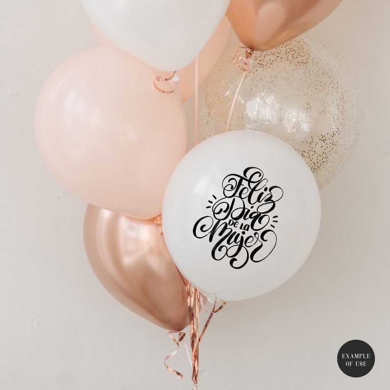 May include: A bunch of balloons in white, peach, and rose gold with a white balloon that says "Feliz Dia de la Mujer" in black calligraphy.