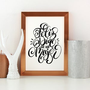 May include: A framed print with the text "Feliz Dia de la Mujer" in black calligraphy on a white background. The frame is copper coloured and is sitting on a white surface with a white antler figurine and two candles.