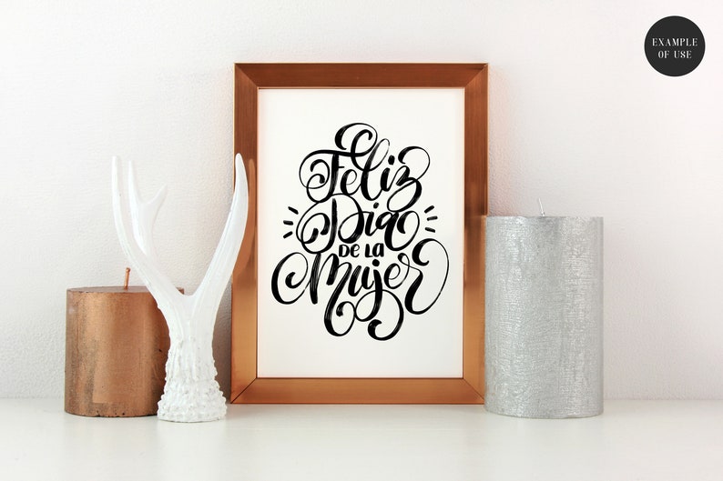 May include: A framed print with the text "Feliz Dia de la Mujer" in black calligraphy on a white background. The frame is copper coloured and is sitting on a white surface with a white antler figurine and two candles.